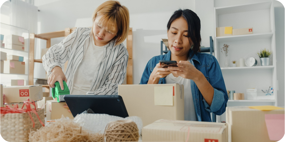 Two women business owners, the woman on the left is preparing some delivery boxes. The woman on the right is handling business on her phone, with a tablet in front of her.